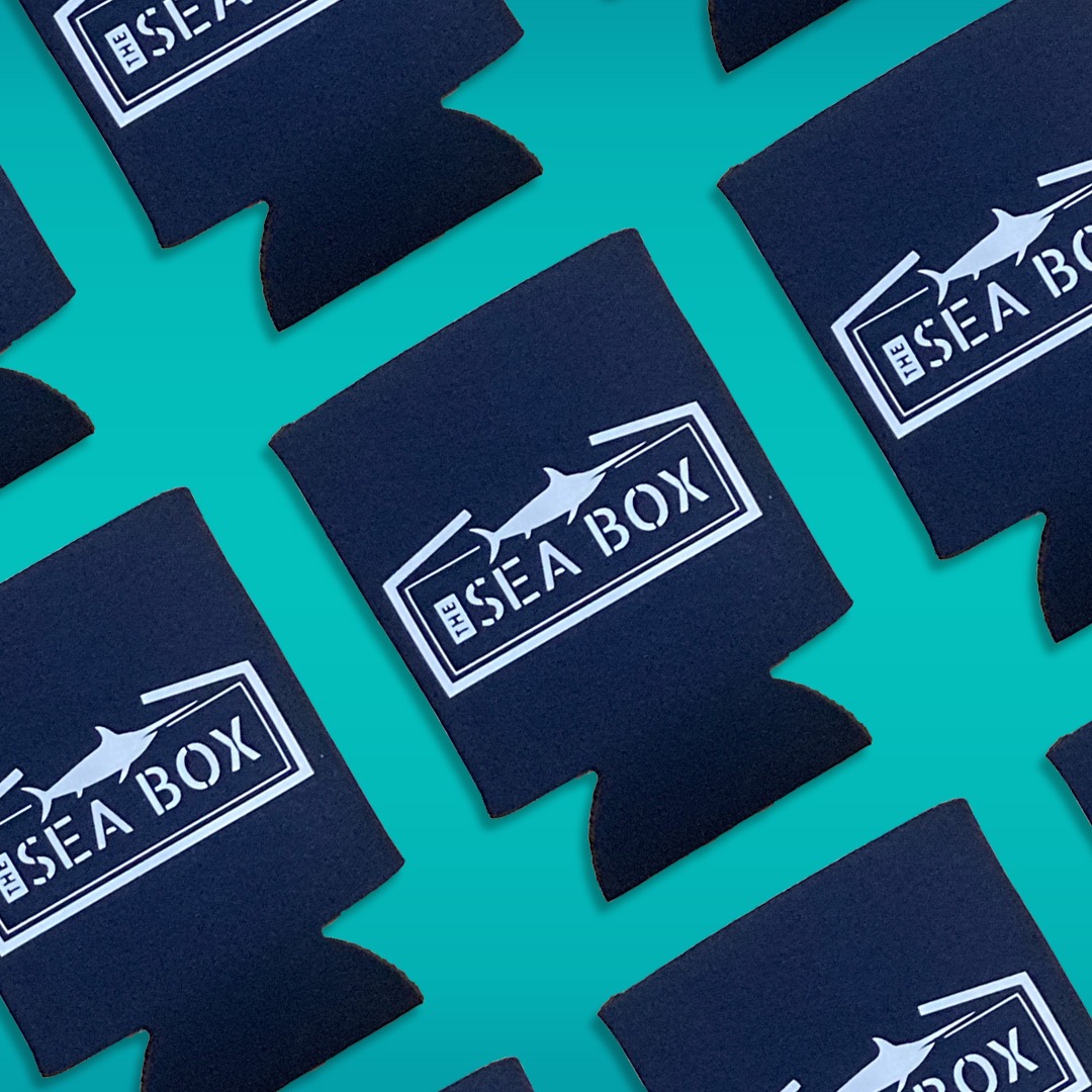 The Sea Box branded beverage koozies featuring a white nautical logo and swordfish illustration on navy blue fabric against a turquoise background.