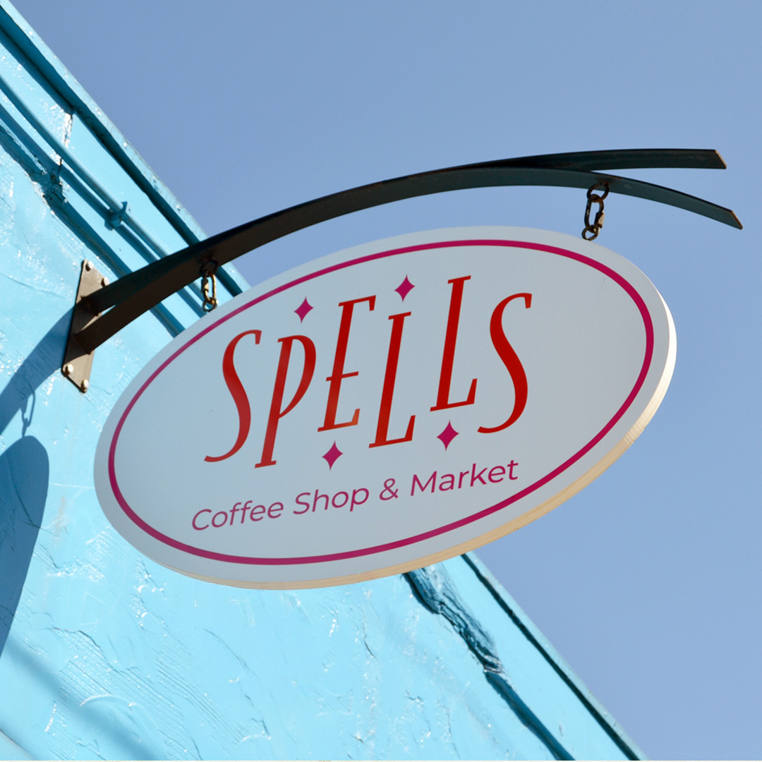 Spells Spells Coffee Shop & Market exterior sign featuring custom orange lettering, pink diamond accents, and an oval white sign mounted on a blue building.